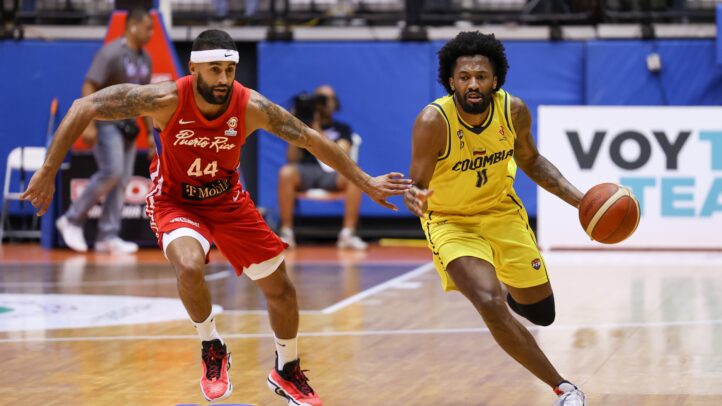 Dos jugadores de baloncesto compiten en la cancha, uno con balón y otro persiguiéndolo.