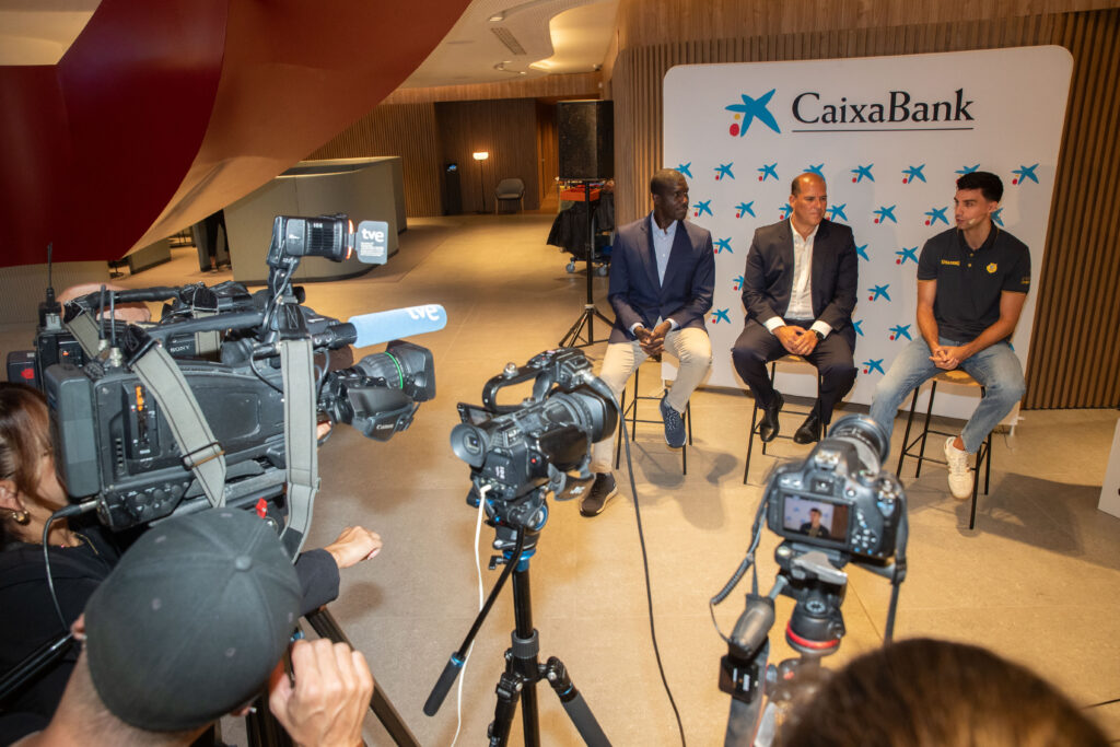Tres personas sentadas en una mesa, rodeadas de cámaras en un evento de CaixaBank.