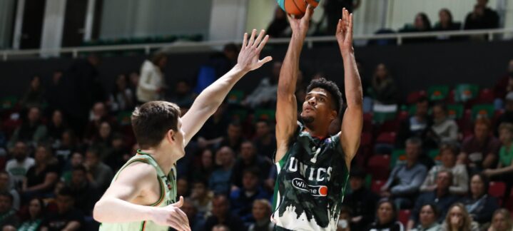 Jugador lanzando a canasta mientras es defendido por un oponente en un partido de baloncesto.