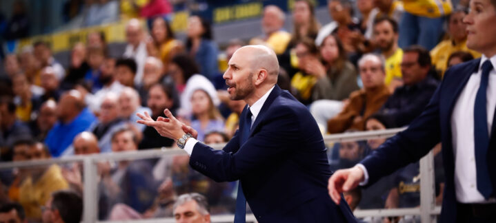 Entrenador dirigiendo al equipo durante un partido, con afición en las gradas.
