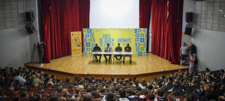 Auditorio con niños aplaudiendo y dos personas en una mesa en el escenario.