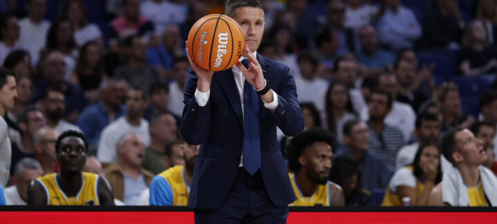 Entrenador sosteniendo un balón de baloncesto y dirigiendo al equipo desde la línea lateral.